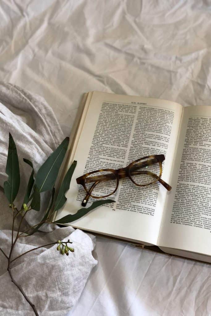 a book on a bed sheet with reading glasses folded on top